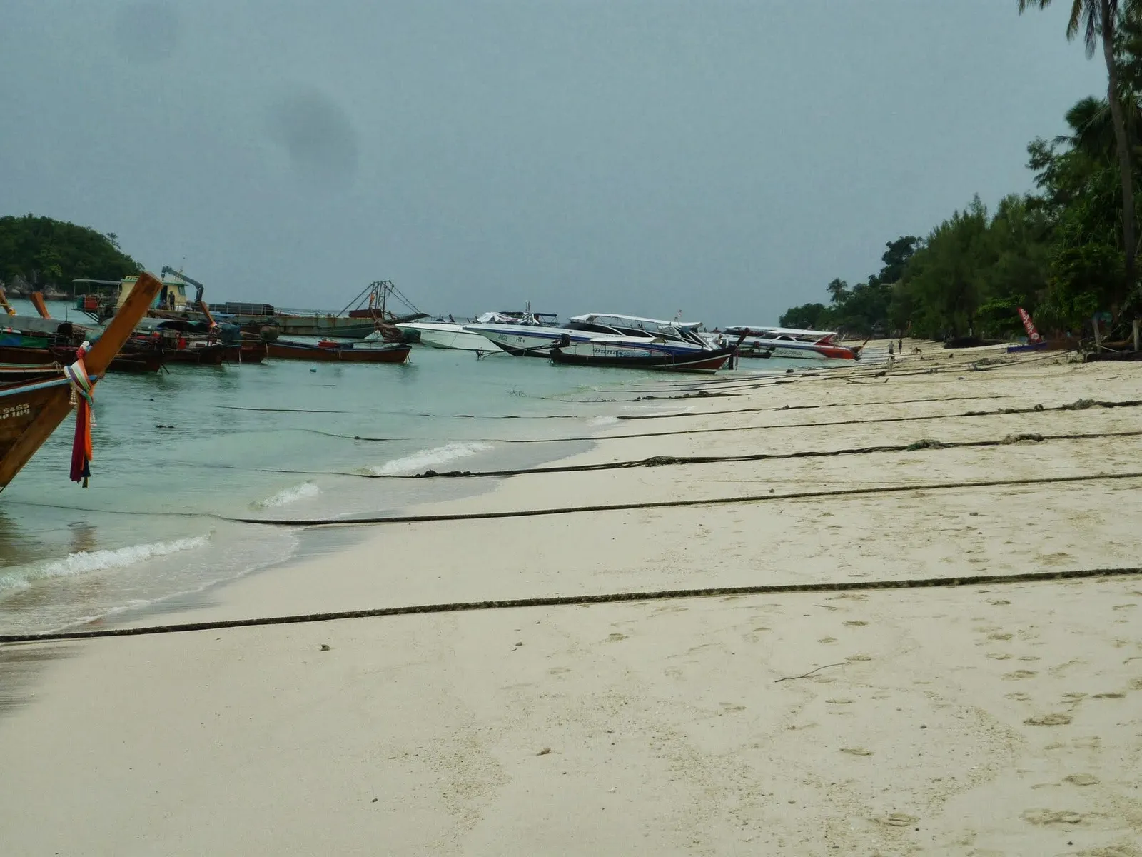 Longtail boats and speedboats moored along a Koh Lipe beach on an overcast day